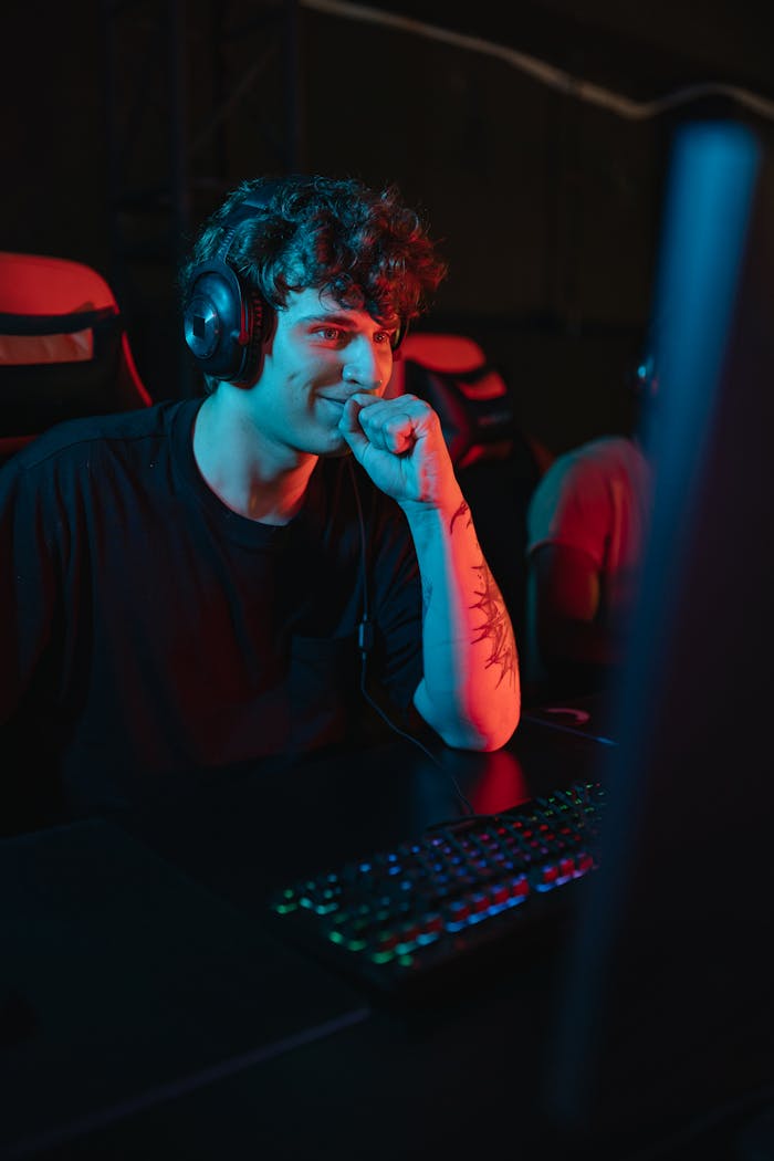 A young man sitting and gaming indoors, wearing a headset with a focused expression under colorful lighting.